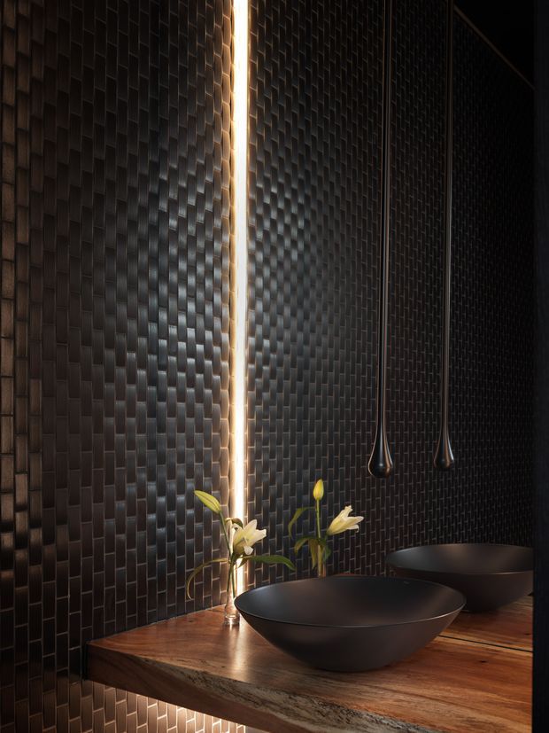 A dramatic, moody bathroom vanity featuring a dark textured tile wall, a floating timber benchtop, two matte black vessel basins, slim black pendant lights, and a narrow vertical strip of warm illumination. A small floral arrangement adds a subtle natural accent.
