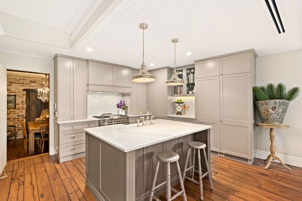 Bright, refined traditional kitchen with a large central island, pale grey cabinetry, white stone benchtop, integrated appliances, pendant lighting, timber flooring, and three stools. A brick-lined dining area is visible through the doorway.