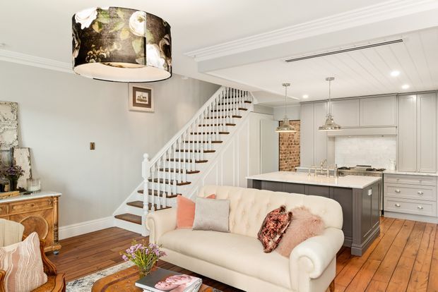 Bright open-plan living room with a cream tufted sofa, timber flooring, decorative pendant lighting, and a white-painted staircase leading toward a contemporary kitchen. The kitchen features grey cabinetry, a central island with a dark benchtop, white splashback, stainless appliances, and an exposed brick accent wall. Traditional architectural detailing is combined with contemporary furnishings.