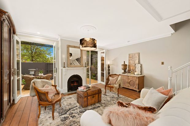 Bright, elegant living room with traditional architectural detailing, timber floors, ornate ceiling cornices, a decorative fireplace, and a white-painted staircase. The room is furnished with a white sofa, vintage timber armchairs, an antique-style carved sideboard, trunk coffee table, patterned rug, and decorative accents. French doors open to a small balcony or terrace, while a mirror reflects the staircase and entry area.