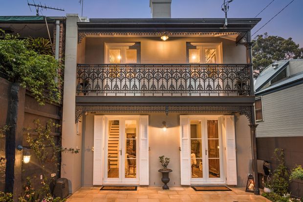 Two-storey attached residential terrace facade at dusk, featuring a warm grey rendered exterior, ornate black wrought-iron balcony balustrade, decorative cornice detailing, white louvred shutters and French doors opening onto a tiled courtyard. Warm interior lighting is visible through the windows and doors, with neighbouring attached homes on either side.