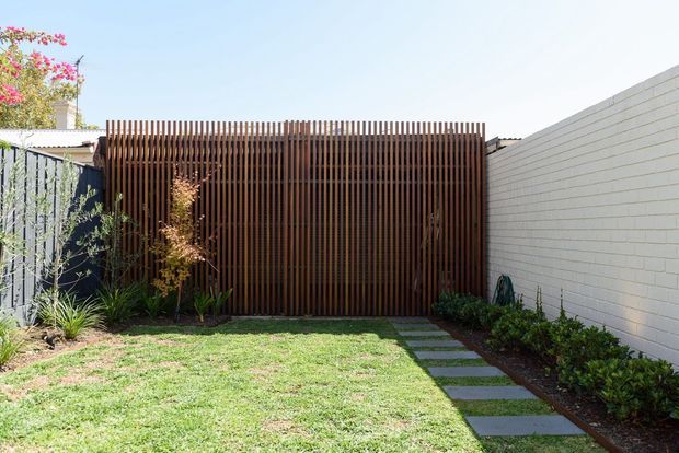 A sunny residential courtyard or backyard enclosed by a tall vertical timber privacy screen and a pale painted masonry wall. A narrow stepping-stone path runs along the wall through a simple lawn and landscaped planting, with a small gate integrated into the timber screen. The setting has a clean, contemporary outdoor design with warm timber contrasted against white masonry and green planting.