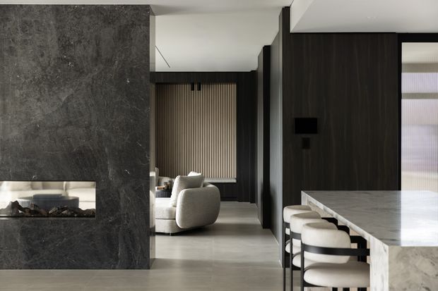 A refined contemporary open-plan interior featuring a dark stone fireplace wall, dark timber wall panelling, a light stone kitchen island with upholstered stools, and a lounge area beyond. Vertical timber screening and restrained neutral furnishings create a sophisticated, high-contrast scheme.