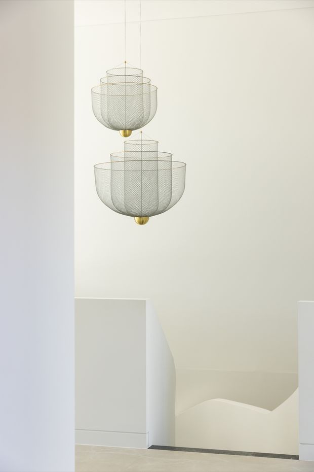 Minimalist interior view of a stairwell featuring two suspended, tiered wire-mesh pendant lights with small gold-toned spherical details. The space has smooth white walls, a sculptural white stair balustrade, pale flooring, and a dark narrow stair-edge trim. The composition is clean, airy, and architectural.