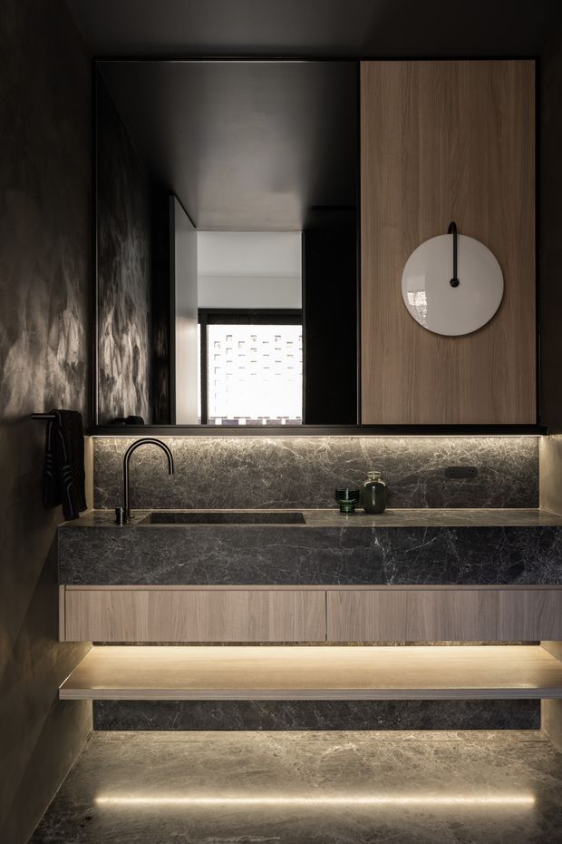 A dark, highly refined compact bathroom vanity with a floating timber-look cabinet, dark veined stone benchtop and integrated backsplash, under-cabinet and toe-kick lighting, black wall finishes, and a round wall-mounted mirror. A black gooseneck tap and undermount basin sit below a large mirror, with a softly lit stone floor and recessed open shelving creating a dramatic, luxurious atmosphere.