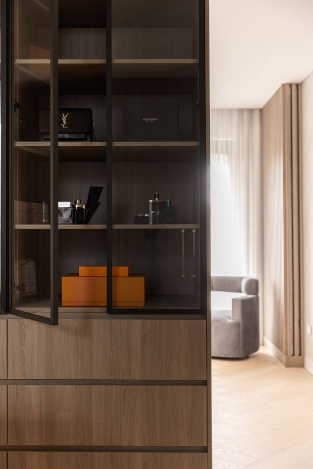 Close-up interior view of a dark timber built-in display cabinet with black-framed glass doors, open shelving, and lower wood drawers. The shelves contain handbags, boxes, and small decorative objects. Through the opening at right, a bright adjoining living area is visible with pale flooring, sheer curtains, vertical timber wall slats, and a rounded upholstered chair.