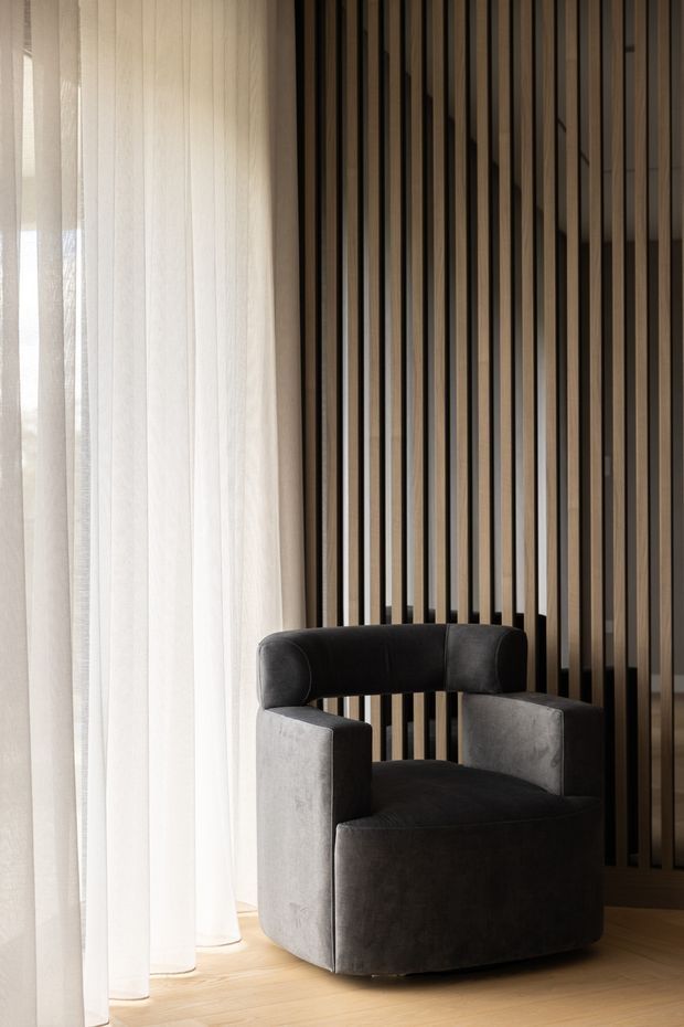 A refined interior corner featuring a sculptural dark grey upholstered armchair positioned beside full-height sheer white curtains. Behind the chair is a dramatic vertical timber slat screen with dark recessed gaps, and the floor is pale timber. The composition is minimalist and contemporary, with soft natural light and warm neutral tones.