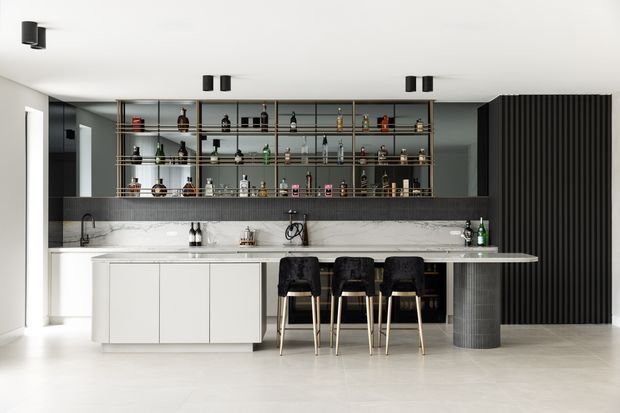 Contemporary home bar and entertaining space with a long white stone island and integrated seating, backed by open metal-and-glass bottle shelving and a marble-look splashback. Dark ribbed wall panelling, black fixtures, and black upholstered bar stools create contrast against the pale floor and cabinetry.