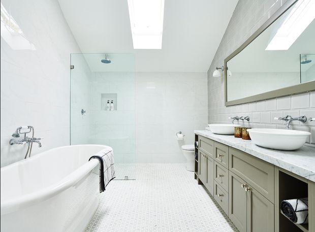Bright, spacious bathroom with a freestanding white bathtub, glass-enclosed walk-in shower, wall-mounted toilet, double vessel basins, and a long grey-green vanity. White tiled walls and small-format mosaic floor tiles create a clean, refined look, with skylights and a large framed mirror adding light and openness.