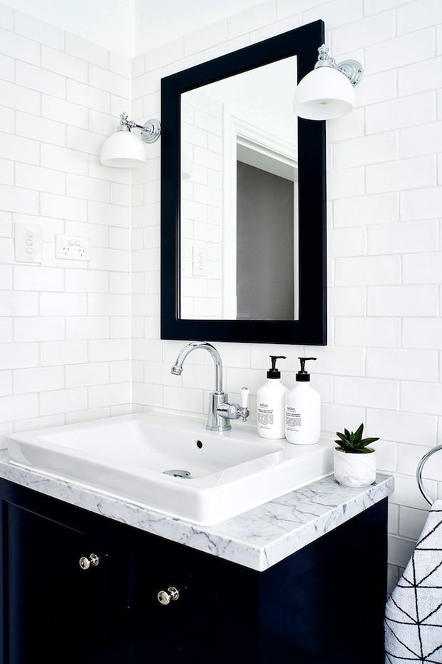 Bright, compact bathroom vanity featuring white subway-tile walls, a white integrated basin, marble-look countertop, dark navy or black cabinetry, chrome tapware, and a large black-framed mirror with matching wall sconces. A small potted plant and toiletries sit on the vanity.