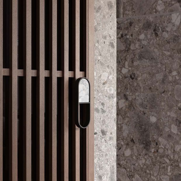 Close-up architectural detail of a dark vertical timber slat screen or door with a distinctive oval black-and-glass handle, set against a textured grey stone wall and a lighter stone edge. The composition highlights rich material contrast and refined contemporary detailing.