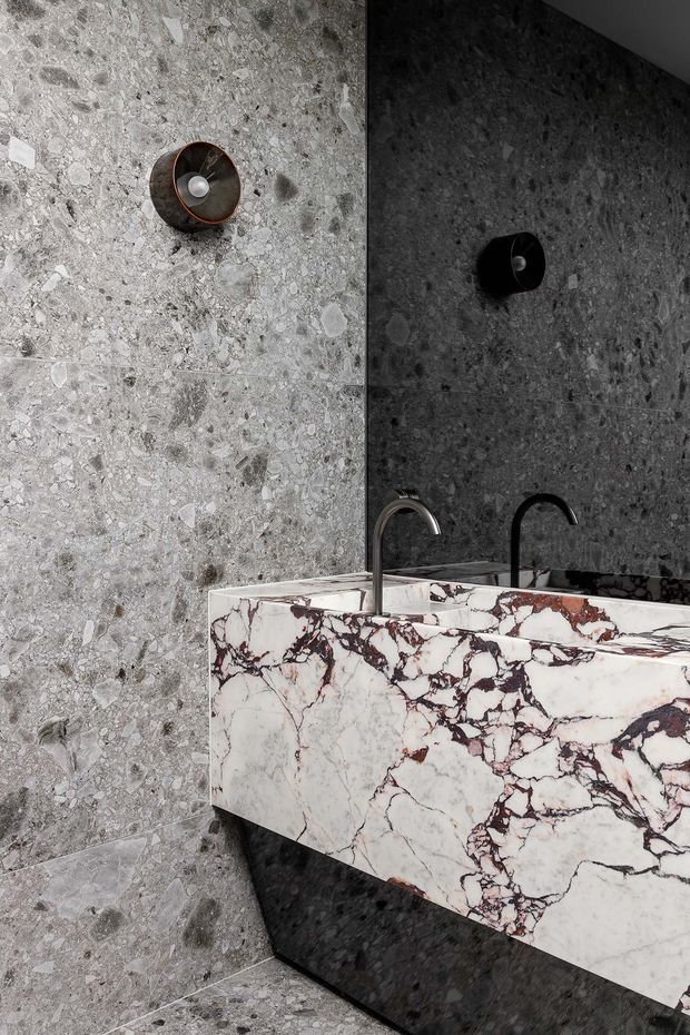 A dramatic contemporary bathroom featuring a floating double vanity with a heavily veined white and burgundy natural-stone benchtop and integrated basin surfaces. The walls and floor are clad in grey terrazzo-like stone with visible aggregate, complemented by dark fixtures, twin curved faucets, and black wall-mounted lighting.