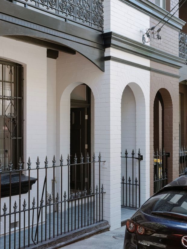 Street-level view of a narrow historic terrace or townhouse facade with white-painted brickwork, repeated arched door openings, dark timber doors, ornate black wrought-iron balcony railings and front fencing, and projecting grey cornices and trim. A parked car occupies the lower right foreground.