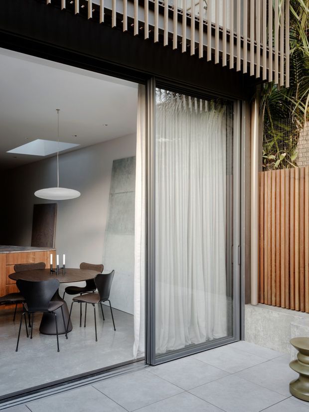 Contemporary indoor-outdoor dining area viewed from a paved terrace through large sliding glass doors. The interior features a round dark dining table with sculptural chairs, timber cabinetry, a minimalist pendant light, polished concrete flooring and a skylight. The facade has dark framing, vertical timber screening and an overhead slatted architectural feature, with greenery visible at the side.
