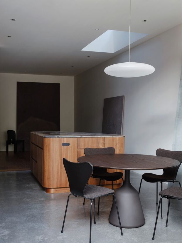 A refined contemporary dining and kitchen space with a large oval dark wood dining table, sculptural pedestal base, upholstered dark chairs, and a compact curved timber kitchen island with a stone benchtop. The room has a polished concrete floor, pale walls, recessed downlights, a large skylight, and a suspended white oval pendant. Dark artwork and furniture create a restrained, moody palette.