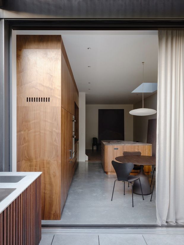 A refined contemporary interior viewed through a wide glazed opening, showing a compact kitchen and dining area. The space features a large warm timber built-in wall and cabinetry, a grey polished concrete floor, a dark stone or composite kitchen benchtop in the foreground, a rounded timber dining table with dark chairs, a sculptural white pendant light, and full-height sheer curtains. Dark-framed glazing and a restrained neutral palette create a clean, minimalist atmosphere.