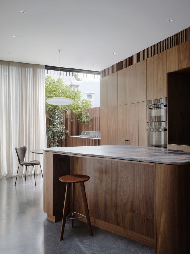 A refined contemporary kitchen with extensive warm timber cabinetry and wall panelling, a large pale stone island with integrated seating, and built-in stainless-steel ovens. The room features polished concrete flooring, a smooth white ceiling with recessed downlights, full-height sheer curtains, and a large glazed opening to a planted courtyard. A slatted timber detail runs along the upper cabinetry and ceiling edge, while a sculptural pendant light and simple stools add understated character.