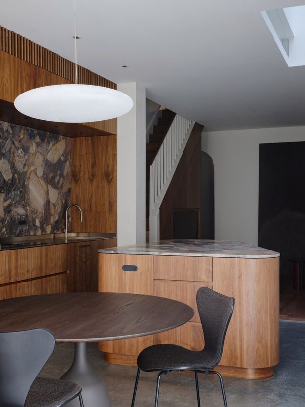 Contemporary open-plan kitchen and dining area with extensive warm timber cabinetry and wall panelling, a dramatic veined stone splashback and island benchtop, dark timber dining table, sculptural pendant light, and a staircase with white balusters leading to an upper level. The space has refined, high-contrast detailing and a partially visible skylight.
