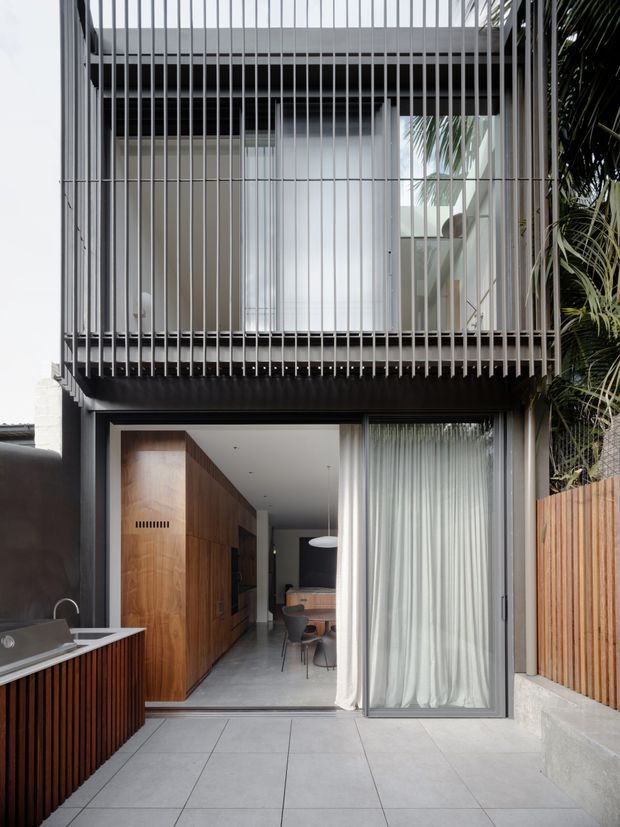 A contemporary multi-level residential courtyard facade with a ground-floor outdoor kitchen and dining area opening into the house. The upper level is screened by closely spaced vertical metal fins, while large glazed openings with sheer curtains connect the interior to the paved courtyard. Dark metal framing contrasts with warm timber cabinetry and a timber privacy fence. Tropical planting is visible at the right edge.