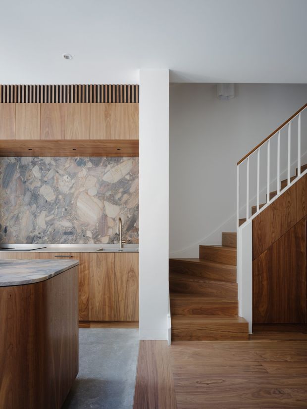A contemporary open-plan kitchen and stairway interior. The kitchen features warm timber cabinetry, a large natural stone backsplash and benchtop, and a dark stone or concrete-like floor. A timber island occupies the foreground. To the right, a timber staircase with white vertical balusters and a timber handrail rises beside built-in timber cabinetry. A white structural column and ceiling create a clean, understated backdrop.