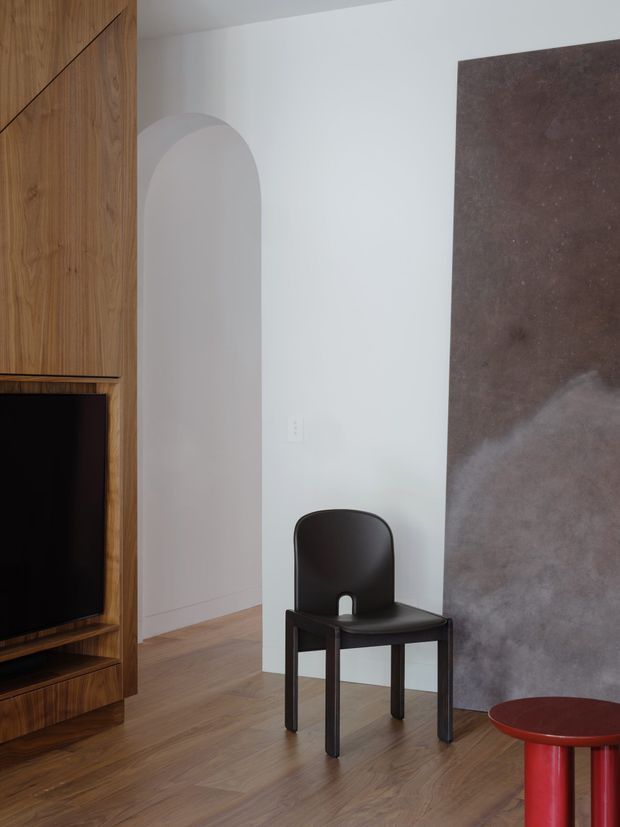 A refined contemporary interior vignette with warm timber built-in cabinetry and media joinery on the left, a white wall with a tall arched opening, and a large dark mottled stone or plaster feature panel on the right. A black sculptural chair and small red side table sit on a warm timber floor.