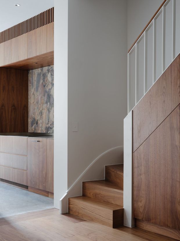 A refined contemporary interior showing a compact kitchen beside a timber staircase. The kitchen features warm wood cabinetry, a dark stone or marble-look splashback, integrated linear ventilation detailing, and a dark benchtop. The staircase has solid timber treads, a white painted stringer and balustrade, and a timber-clad understair wall. Neutral plaster walls and timber flooring create a calm, cohesive palette.