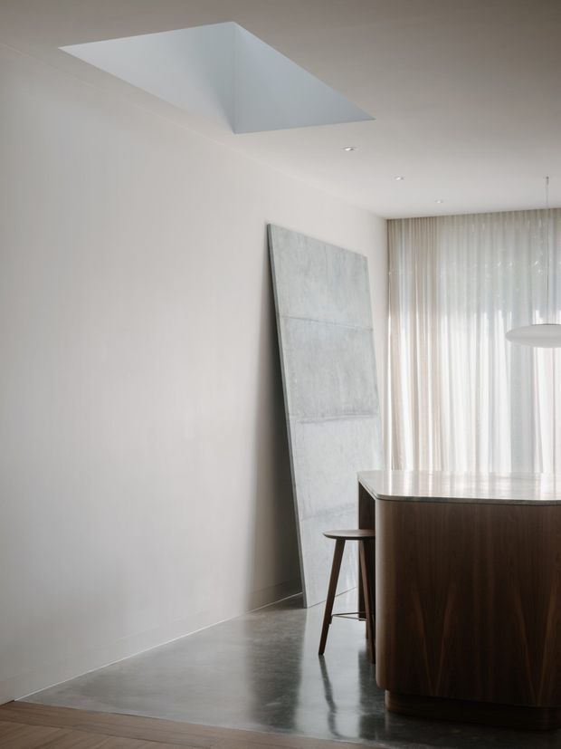 A minimalist dining area with a dark wood island or dining table in the foreground, paired with a simple wooden stool. The room has smooth white walls and ceiling, a polished grey concrete floor, sheer full-height curtains, and a large pale abstract artwork leaning against the wall. A dramatic recessed ceiling opening or skylight adds architectural interest, while a simple white pendant light hangs near the window.