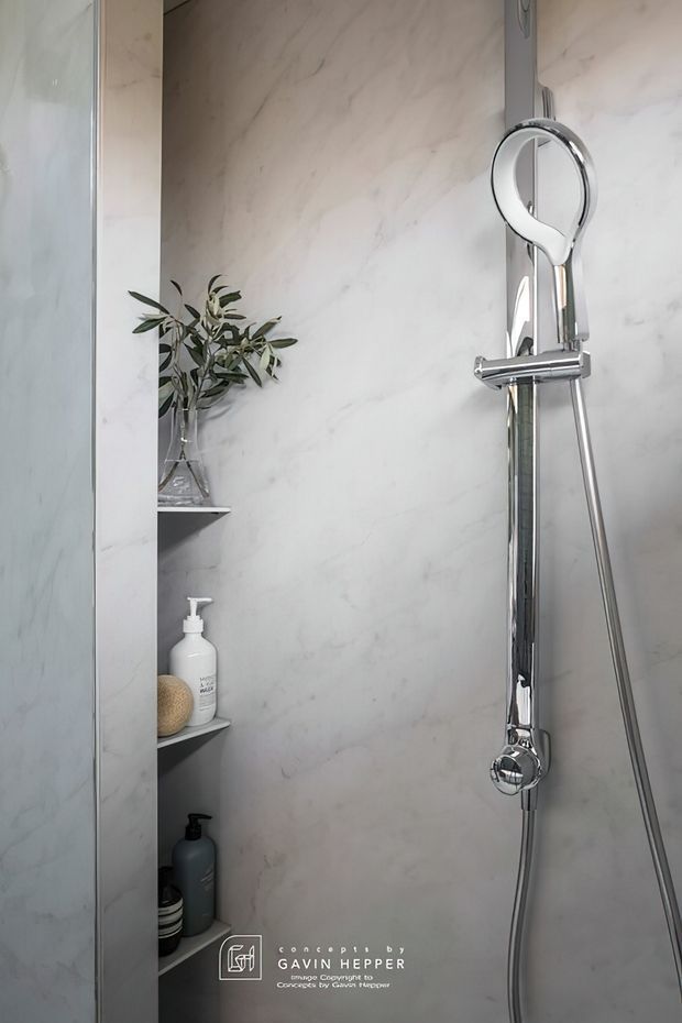 Close-up view of a contemporary shower enclosure with large pale marble-look wall panels, recessed open shelving holding toiletries and decorative greenery, and a polished chrome handheld shower rail with circular control fixture. A glass shower screen is visible at the left edge.
