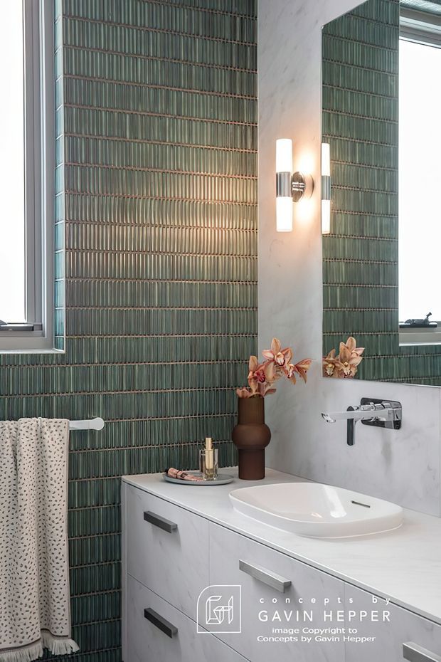 A contemporary bathroom vanity area featuring a white integrated basin and wall-mounted chrome tapware. The walls are finished with vertically ribbed green-grey tiles, complemented by a white marble-look wall surface, a large mirror, modern wall sconces, and a dark decorative vase with flowers. A towel rail and patterned towel are visible beside the vanity.