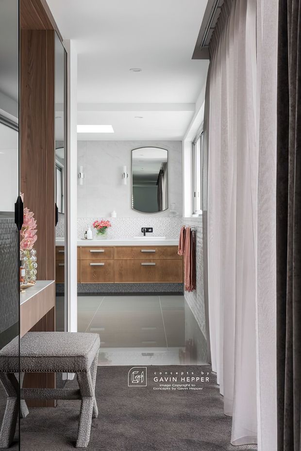 A refined contemporary bathroom viewed from an adjoining dressing or bedroom area. The room features a floating warm timber vanity with integrated drawers, a white stone or marble-look benchtop, pale mosaic backsplash, large grey floor tiles, a rounded rectangular mirror, wall sconces, and full-height windows dressed with sheer and dark curtains. A built-in timber and mirrored joinery wall and upholstered bench are visible in the foreground.