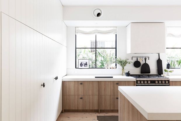 Bright contemporary kitchen with white vertical-panel cabinetry, warm wood-grain lower cabinets, a large white island, black-framed windows, integrated appliances, and black cooking accessories displayed against a white splashback. Natural light and indoor greenery create a clean, warm atmosphere.