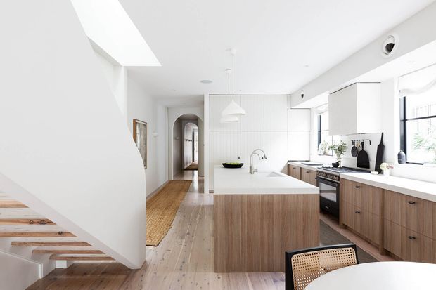 Bright contemporary open-plan kitchen viewed from beside a floating timber staircase. The room features a large central island with white benchtop and vertically grained timber cabinetry, matching timber kitchen cabinetry, white tiled or panelled walls, pale timber flooring, white pendant lighting, integrated appliances and generous windows. A hallway and adjacent living or dining area are visible beyond the kitchen.