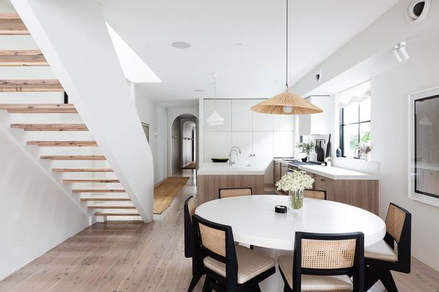 Bright open-plan kitchen and dining area with a round white dining table, black-framed cane dining chairs, light timber cabinetry, white benchtops, pale timber flooring, and a floating timber staircase. A skylight, pendant lighting, arched hallway opening, and large windows contribute to the airy contemporary interior.
