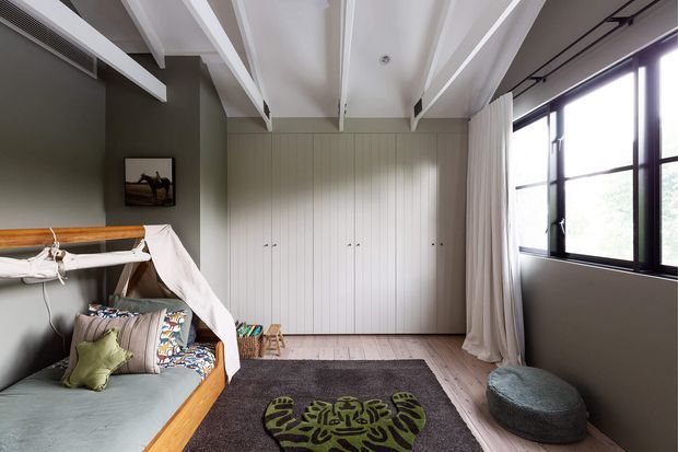 A bright, contemporary child's bedroom with a timber-framed daybed featuring a draped canopy, patterned cushions, and a soft green throw. The room has muted olive-grey walls, full-height white vertical-panel built-in cabinetry, pale timber flooring, a dark rug, exposed white ceiling beams, large black-framed windows, and a low upholstered floor cushion.