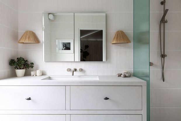 Bright, refined bathroom vanity with a wide white floating-style cabinet, integrated basin and wall-mounted taps. Pale vertical wall tiles create a clean backdrop, while a mirrored cabinet, woven wall sconces, small plants and decorative objects add warmth. A glass-enclosed shower with exposed shower fittings is visible on the right.