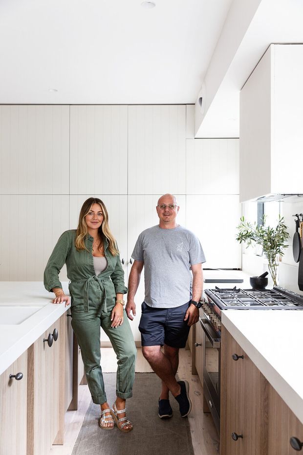 Bright, narrow contemporary kitchen with white vertical-panel cabinetry extending across the walls, pale timber lower cabinets, and light stone benchtops. A large central island with dark knobs occupies the foreground, while a gas cooktop, stainless rangehood, and open shelving sit along the right-hand wall. Two people stand in the kitchen, providing scale. The space has a clean, minimal, light-filled appearance.