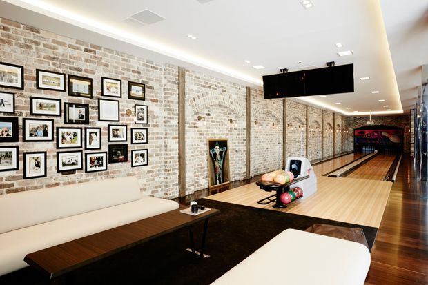 A spacious indoor bowling and entertainment area with exposed light brick walls, arched brick niches, framed artwork, polished timber flooring, dark carpet, and contemporary cream seating. Bowling lanes extend along the right side, with a billiards or lounge area in the foreground and modern recessed lighting overhead.
