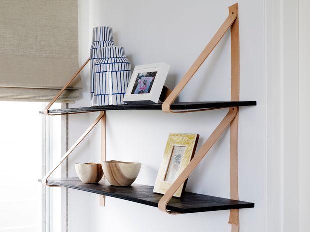 Close-up of a minimalist wall-mounted shelving system with black horizontal shelves supported by distinctive light timber strap-like brackets. The shelves display a tall blue-and-white patterned ceramic vase, framed photographs, wooden bowls, and a framed artwork. The setting is bright and neutral, with a window blind visible at left.