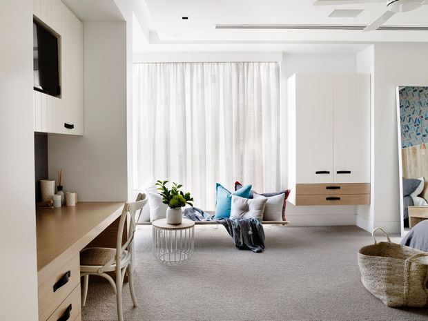 Bright, refined bedroom with a built-in timber desk and shelving, upholstered seating area, large curtained window, integrated storage cabinetry, and soft neutral finishes. A mirror reflects part of the bed and adjacent furnishings.