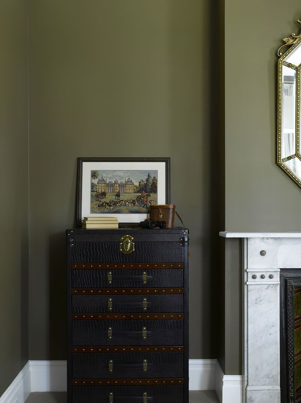 A refined interior vignette with an olive-grey painted wall, white skirting, a dark crocodile-textured leather chest of drawers with brass hardware, and a white marble fireplace partially visible at right. A framed architectural artwork, books, and a small decorative object sit atop the chest, while an ornate gold-framed mirror is cropped at the upper right.