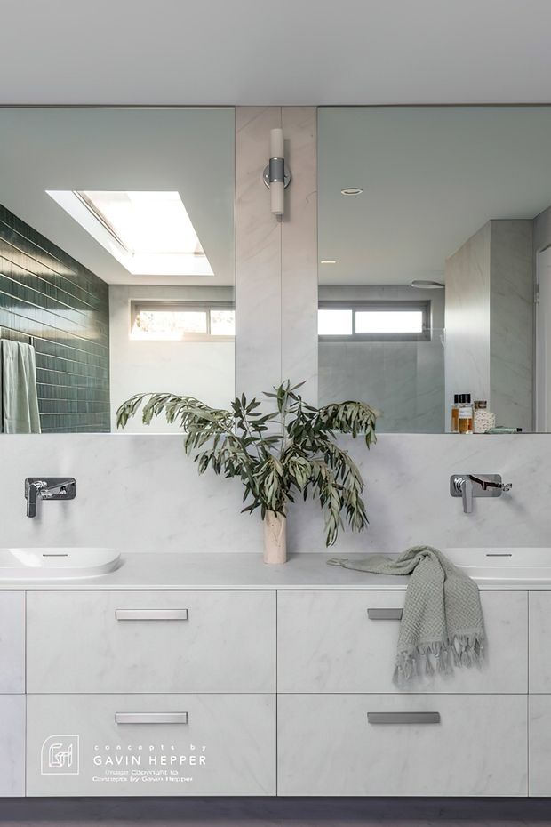 Contemporary bathroom vanity with a double basin, wall-mounted chrome tapware, pale marble-look cabinetry and splash surfaces, a large mirror, skylight, clerestory windows, and a dark green textured tile feature wall. A potted branch and folded towel add restrained natural accents.