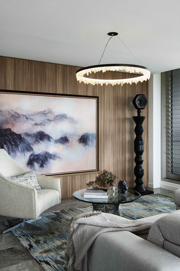 A refined contemporary living room featuring a large abstract artwork, vertical timber wall panelling, a sculptural black floor lamp, round glass coffee table, upholstered lounge seating, and a statement circular pendant light. Layered neutral furnishings and a patterned area rug create a sophisticated, warm interior.