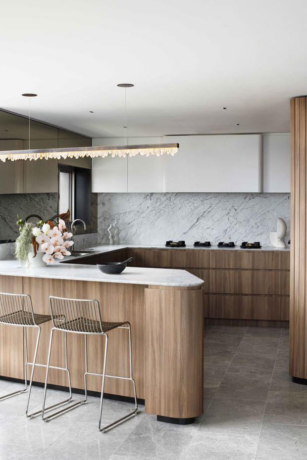 A refined contemporary kitchen with a large rounded island clad in warm vertical-grain timber cabinetry and topped with a pale veined stone benchtop. The rear wall features matching timber drawers, minimalist white overhead cabinetry, and a dramatic veined stone splashback. Grey stone-look flooring, slim metal bar stools, integrated appliances, and a linear pendant light complete the space.