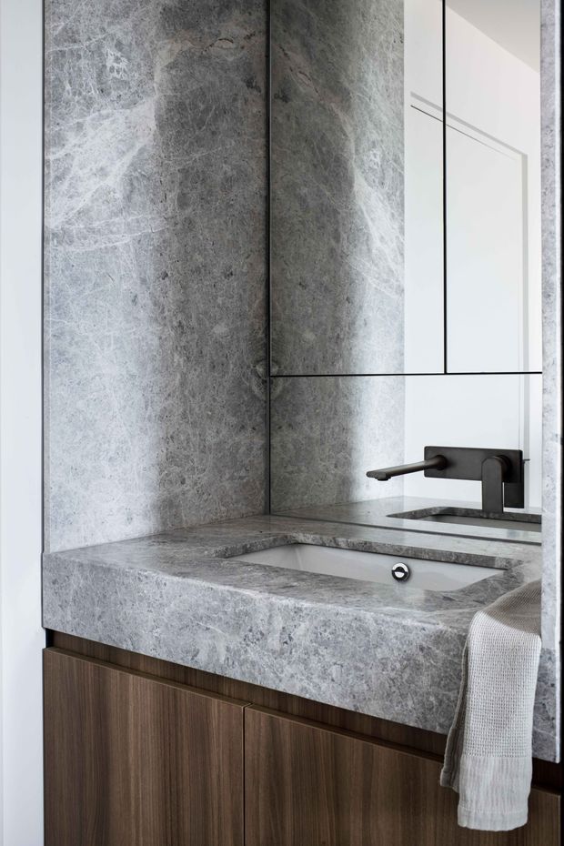Close-up view of a refined bathroom vanity featuring a pale grey veined stone benchtop and integrated rectangular basin, dark wood-grain cabinetry, a wall-mounted dark metal tap, and a large mirrored cabinet above. The space has a restrained contemporary appearance with high-contrast stone, timber, and black metal detailing.