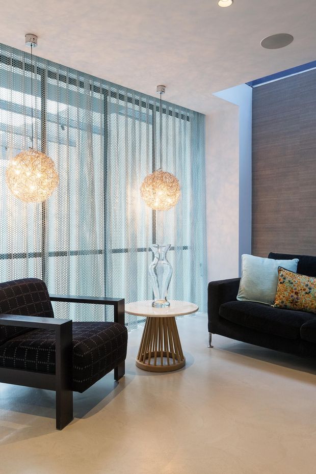 A refined contemporary sitting area with a black upholstered armchair and sofa arranged around a small round side table. Floor-to-ceiling patterned metal mesh curtains filter daylight across the glazed wall, while spherical pendant lights, a textured dark feature wall, pale flooring, and decorative cushions add visual interest.