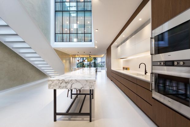 A spacious contemporary kitchen in an open-plan interior, featuring a marble island with dark metal legs, extensive dark timber cabinetry, integrated stainless-steel appliances, a black tap, and a dramatic double-height glazed void above the stairway. White floors, walls and ceilings create a bright, refined atmosphere.