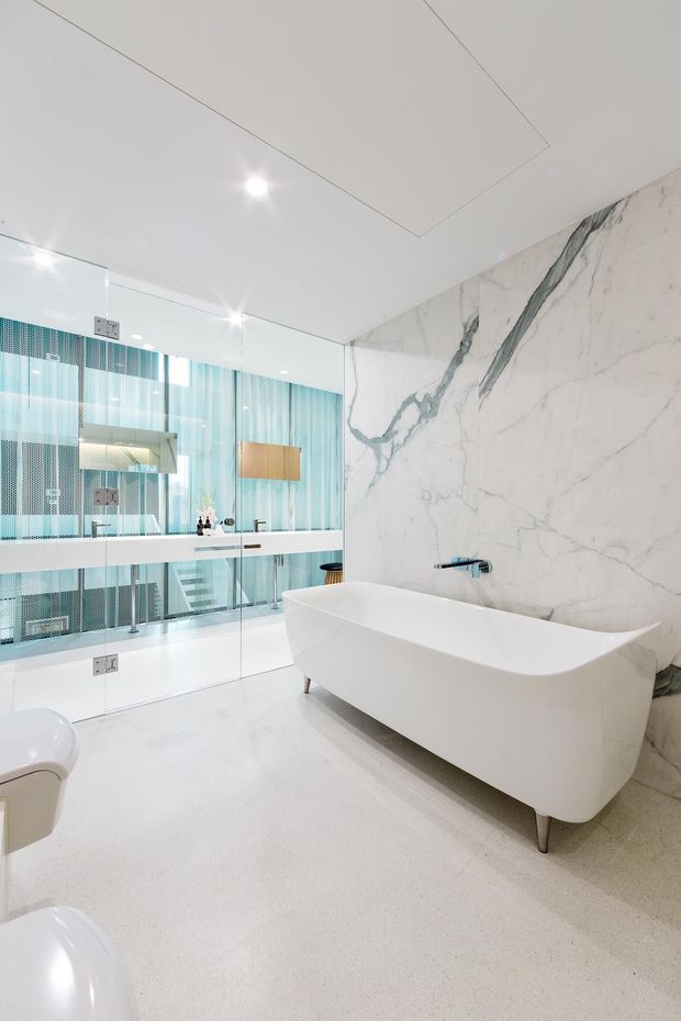 Bright, luxurious contemporary bathroom featuring a freestanding white bathtub, full-height white marble-look wall slab with dramatic grey veining, pale terrazzo-like flooring, frameless glass partition, wall-mounted chrome fixtures, and a turquoise glass-screened area beyond.
