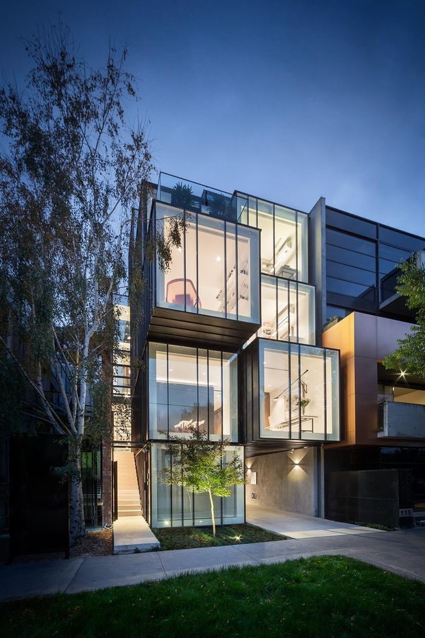 Dusk exterior view of a multi-storey contemporary urban residence with projecting box-like volumes, extensive floor-to-ceiling glazing, dark metal cladding, concrete surfaces, and warm interior lighting. Mature trees and a narrow landscaped frontage frame the building, with a driveway or garage entrance at ground level.