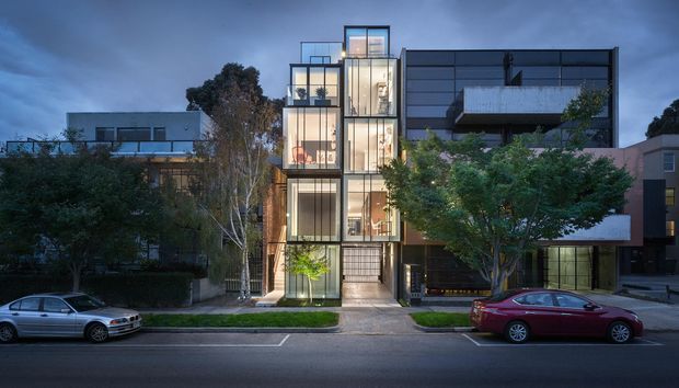 Twilight street view of a contemporary multi-storey urban residential building with a narrow glazed central stair or living volume, dark horizontal cladding, projecting balconies, extensive floor-to-ceiling glazing, and landscaped street frontage.