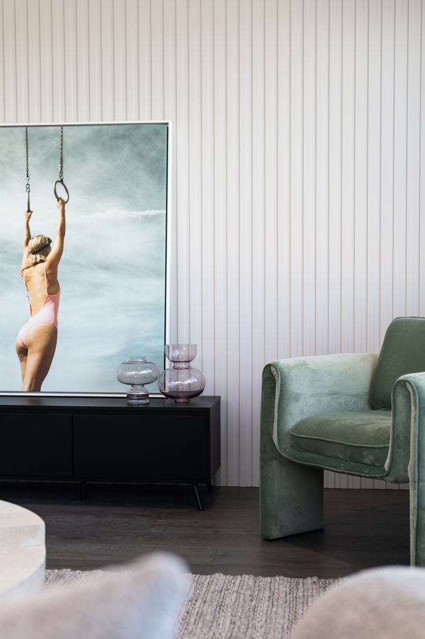 A refined living room corner with a dark timber or black low-profile sideboard, sculptural glass vessels, a large framed artwork or mirror, and a plush sage-green upholstered armchair. The wall is clad in narrow vertical white panels, with dark timber flooring and a textured neutral rug creating a contemporary, softly layered interior.